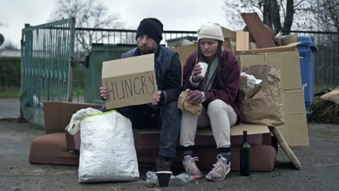 Two dirty and poorly dressed homeless people, a man and a woman, sit by a pile Stock Footage 244119628