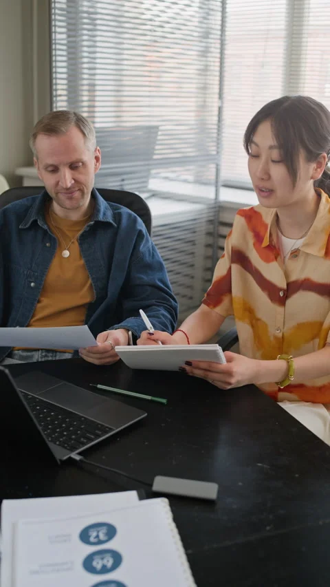 Two Diverse Employees Reviewing Documents at Table with Laptop in Office Stock Footage 335760022