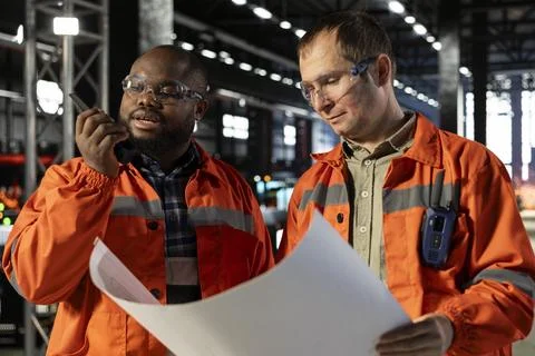 Two diverse engineers use plans to oversee construction and production tasks Stock Photos