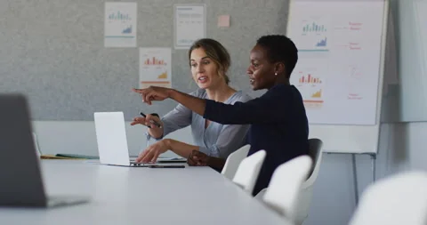 Two diverse female colleagues sitting at desk with laptop and discussing in Stock Footage 154716396