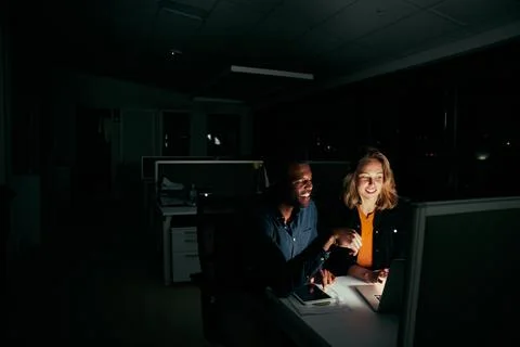 Two diverse happy colleagues working on laptop until night in dark office Stock Photos