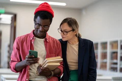 Two diverse students standing in classroom using smartphone during study break Stock Photos