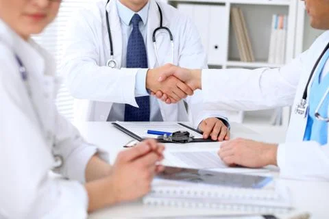 Two doctors shaking hands to each other sitting at the table in hospital office Stockfoto's