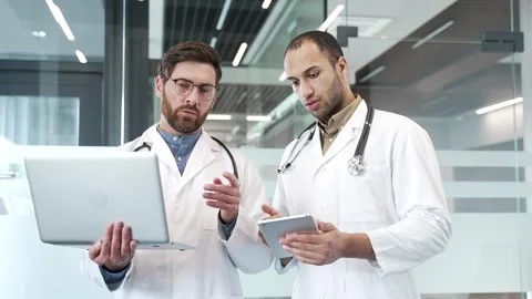 Two doctors talking over laptop and tablet standing in hospital office. Medical  Stock Footage 303413108
