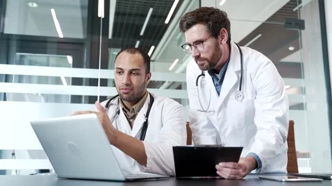Two doctors in white coats collaborate over laptop and folder while sitting  Stock Footage 309644768