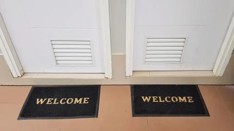 Two doormats lying in front of two doors with welcome written on it Stock Photos