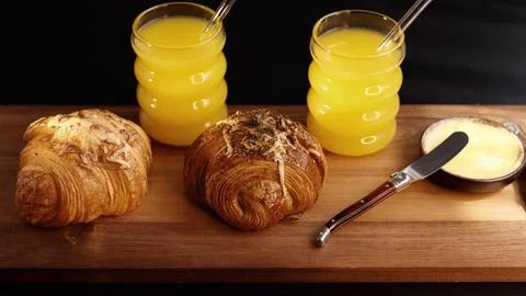 Two drinks with straws beside two croissants, butter placed on wooden board Stock Photos