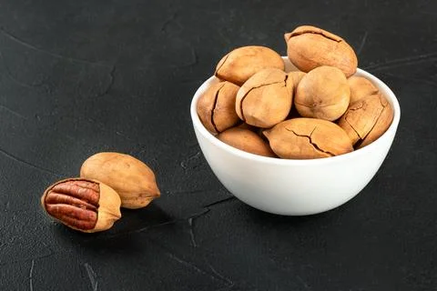 Two dry pecans with a full white bowl of nuts close-up on a dark background Stockfoto's