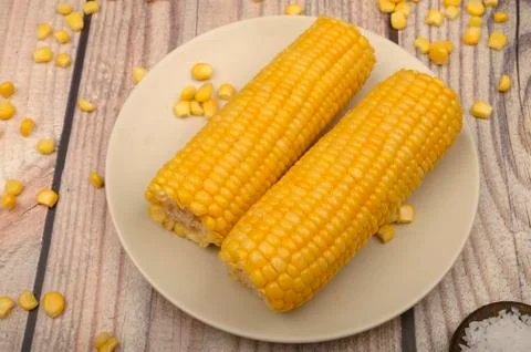 Two ears of boiled corn in a plate, corn kernels and coarse salt on the table Stock Photos