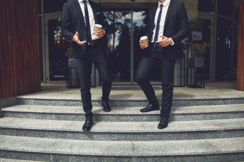 Two elegant men holding cups of coffee while going outdoors Stock Photos