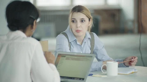 Two employees girlfriends are sitting at the same desk with laptop, chatting and Stock Footage 124328027