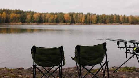 Two empty fishing chairs by calm autumn lake with rods on tripod, peaceful Video stock 320140573