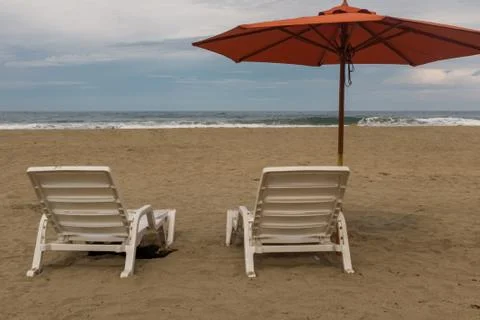 Two empty plastic white chairs under an orange beach umbrella on a deserted b Stock Photos
