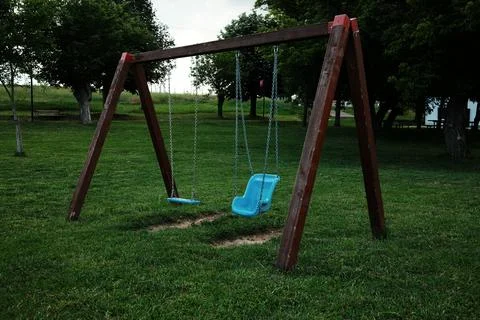Two empty swings, including a toddler seat, hang from a wooden frame in a quiet Stock Photos
