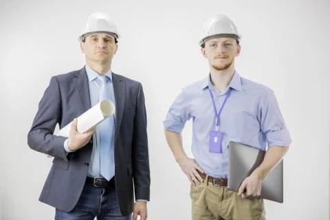 Two engineers confidently look at camera with smile, holding laptop and drawing Stock Photos