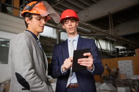 Two engineers discuss a project using a tablet while they are in the factory. Stock Photos