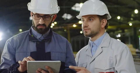 Two engineers in helmets standing in the middle of factory room, scrolling on a Stock Footage 84438670
