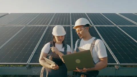 Two engineers in protective gear talk over a laptop in a field of solar panels. Stock Footage 263753706