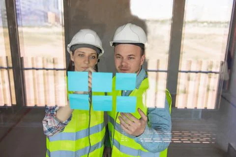 Two engineers in protective vests and white helmets glue stickers on the window Stock Photos