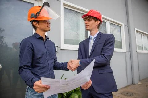 Two engineers shake hands while looking at a floor plan during construction. Stock Photos