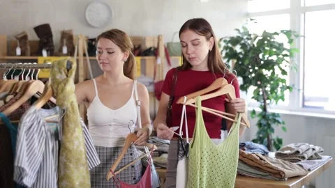Two female friends are standing in the store near the clothes hangers, they are Stock Footage 346125947