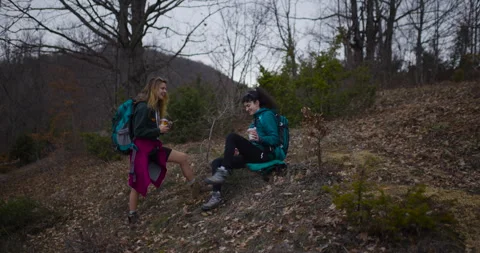 Two Female Friends Take a Break During Their Hike in the Mountains to Enjoy a Stock Footage 338357049