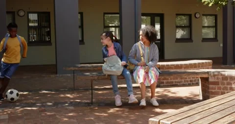 Two female students sitting on bench at school, watching boy entering dribbling Stock Footage 337294213