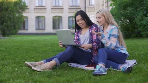 Two female students sitting on grass and choosing new dresses on laptop, device Stock Footage 77849685
