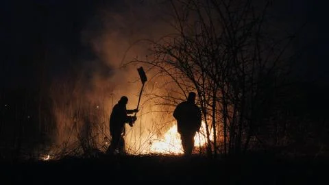 Two firefighters with fire flappers extinguish a fire in the forest at night Stock Photos