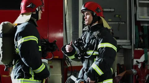 Two firemen in protective uniform and red helmets talk near fire truck Stock Footage 276368485