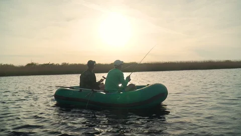 Two fishermen in the rays of the sunset catch fish from an inflatable boat 库存影片 128133099