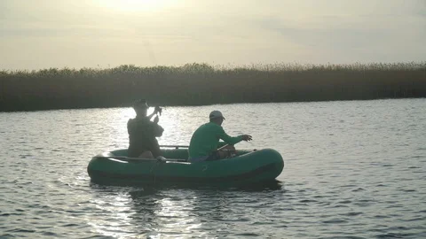 Two fishermen in the rays of the sunset catch fish from an inflatable boat 库存影片 128133410