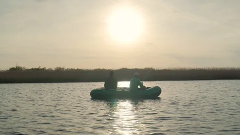Two fishermen in the rays of the sunset catch fish from an inflatable boat 库存影片 128138097