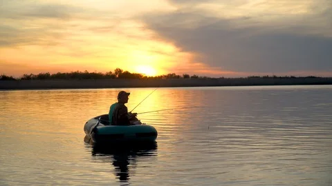 Two fishermen in the rays of the sunset catch fish from an inflatable boat 库存影片 128138306
