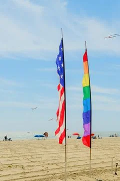 Two flags, an American flag and a rainbow flag, stand on a beach with peopl.. Stock Photos
