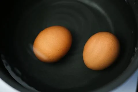 Two fresh brown eggs in a dark saucepan with cold water Stock Photos