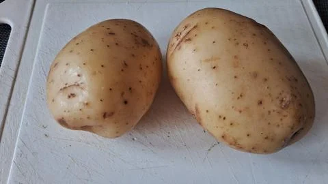 Two fresh raw potatoes sitting on a scratched white plastic board Stock Photos