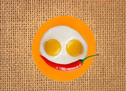 Two fried eggs and red hot chilly pepper on orange plate, knife and fork on b Foto stock