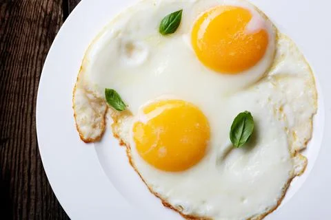Two fried eggs in a white ceramic plate with basil leaves on an old wooden ta Stock Photos