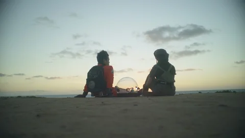 Two friends in astronauts suits getting up and walking away on beach at sunset Stock Footage 108965305