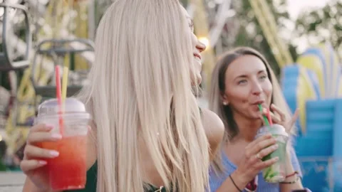 Two friends dancing colorful drinks at an amusement park during a sunny Stock Footage 312780070