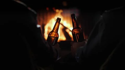 Two friends holding beer bottles while sitting in front of a cozy fireplace.. Stock-Footage 334606080