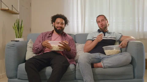 Two friends joyfully share popcorn while watching their favorite TV show in a Stock Footage 305494290
