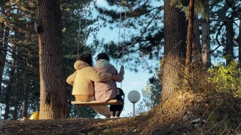 Two friends sharing a moment on a swing amid tall pine trees at sunset Video stock 311805729