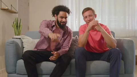 Two friends sit side by side on a cozy couch, comfortably using their Stock Footage 305822119