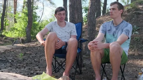 Two friends sitting on chairs in a pine forest and talking Stockbeeldmateriaal 135080171