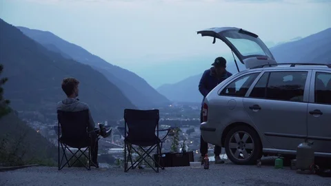 Two friends sitting down in deck chairs besides car, toasting with a beer, 4K Stock Footage 129614670