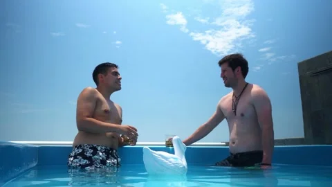 Two friends talking and drinking whiskey in glass glasses inside a pool on a Vidéo 173506299