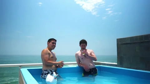 Two friends toasting with whiskey inside a pool on a terrace with a beautiful Stock Footage 173506228