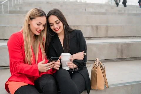 Two friends using their mobile phone while sitting outdoors. Stock Photos